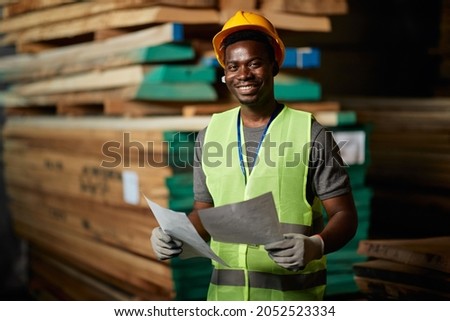 Happy African American worker going through delivery plans at lumber distribution warehouse and looking at camera. Royalty-Free Stock Photo #2052523334