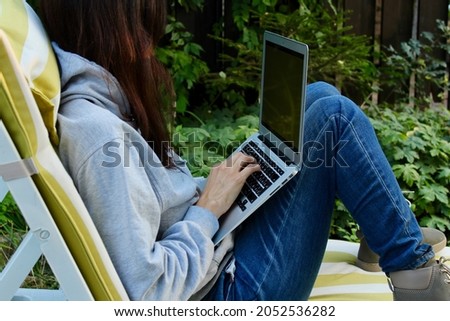 Young lady with red hair working on a silver laptop with green screen wearing grey hoodie and jeans and sitting in a chaise lounge in a green garden. Freelancer freelance job work from home concept