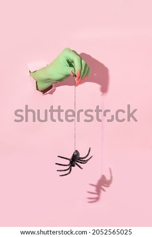 Halloween creative layout with green witch hand with bright pink nails holding spiders web with spider against pastel pink background. Aesthetic holiday season  idea. Minimal Halloween concept.