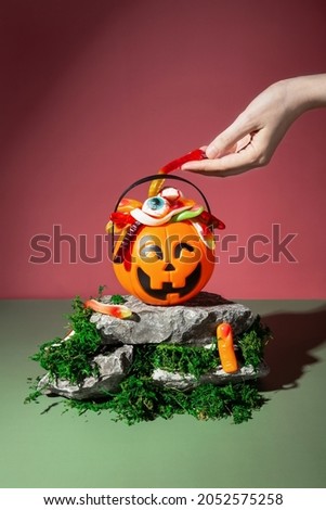 Funny Halloween scenery. Trick or treat. Pumpkin Jack filled with various creepy sweets stands on stones and moss. Female hand pulls out jelly worm