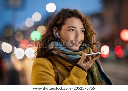 Happy woman recording message using voice on smartphone. Happy woman recording audio on mobile phone in the street at evening. Smiling woman speaking on her smart phone in winter evening. Royalty-Free Stock Photo #2052575765