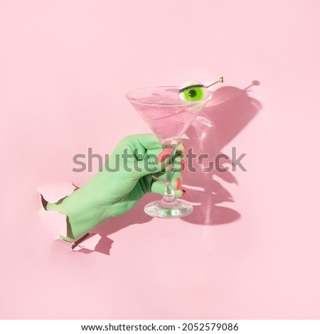 Halloween creative layout with green witch hand with bright pink nails holding martini cocktail glass with eyeball against pastel pink background. Aesthetic season idea. Minimal Halloween concept.