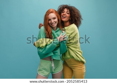 Young african woman and european woman in friendly embrace with copy space. They are wearing light green hoodie. Red-haired lady and brunette spend time together. Lifestyle, female beauty concept