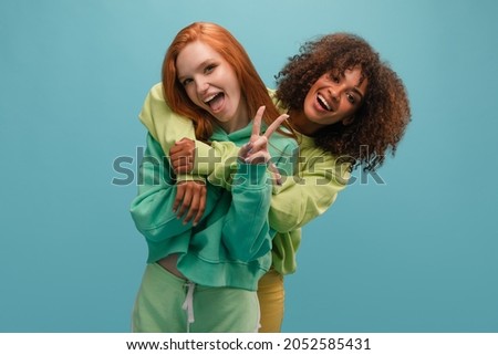 Dark-skinned girl hugs her fair-skinned friend from behind on blue background in studio. They have smile with open mouth. Redhead shows peace with her left hand.
