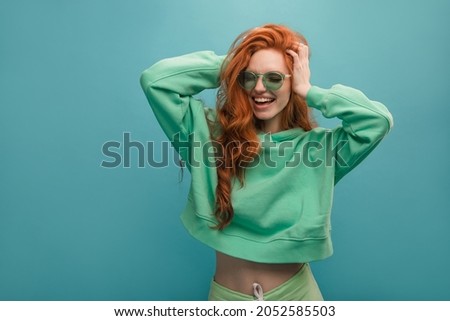 Disheveled young woman of European appearance is holding her head against blue background. Red-haired hipster in translucent glasses is wearing green hoodie. Concept of good mood, emotion Royalty-Free Stock Photo #2052585503