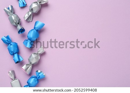 Many candies in light blue and silver wrappers on violet background, flat lay. Space for text