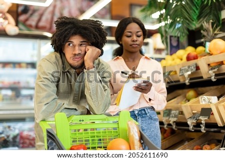 Grocery Shopping. Bored African Husband Standing With Shop Cart While Wife Taking Notes In Shop List Buying Food In Supermarket. Family Budget And Household, Financial Crisis. Selective Focus Royalty-Free Stock Photo #2052611690