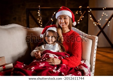 Winter Holidays. Portrait of cheerful woman and small girl in Santa Claus hats celebrating Xmas evening and New year at home, sitting on the couch under cozy warm red blanket, watching tv cartoons