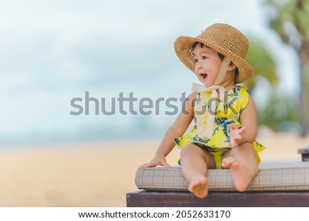 A baby, in a bright yellow dress, sits on a beach chair and wears a hat on a family vacation, excited and enjoying the sea. Royalty-Free Stock Photo #2052633170