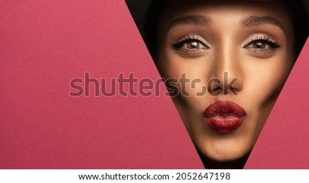 Face of a young beautiful girl with a bright make-up and with plump red lips peeks into a hole in pink paper.