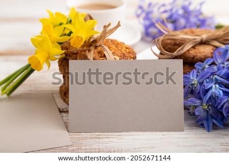 Gray paper business card mockup with oatmeal cookies with spring snowdrop flowers bluebells, narcissus and cup of coffee on white wooden background. side view, close up, still life.