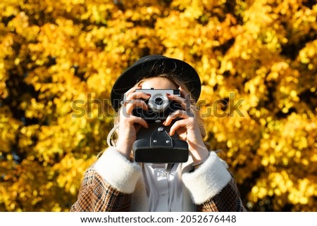 Portrait of young woman hipster photographer in coat and hat taking photo on retro film vintage camera in autumn park outdoors. Selective focus on photographic equipment. Hobby and lifestyle.
