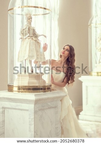 Beautiful woman with long hair in a light dress and a big doll ballerina in the theater under a glass dome