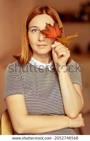 Portrait of a young woman covering one eye with an autumn fallen maple leaf. Autumn mood.