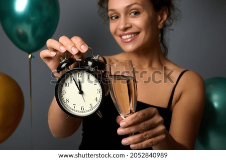 Close-up of an alarm clock and flute of champagne in the hands of blurred smiling beautiful woman, isolated over grey background and inflated green metallic and golden air balloons, copy space for ad