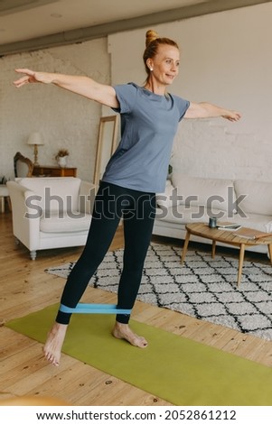 Smiling fit woman in sport clothes keeping body in good shape, having indoor training using resistance loop band, doing leg lifts, holding arms out of the side trying to catch balance