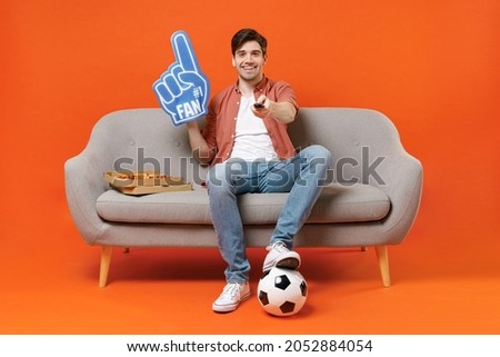 Young man football fan in shirt support team with soccer ball sit on home sofa watching tv live stream hold pizza foam glove finger switch channel isolated on orange background People sport concept