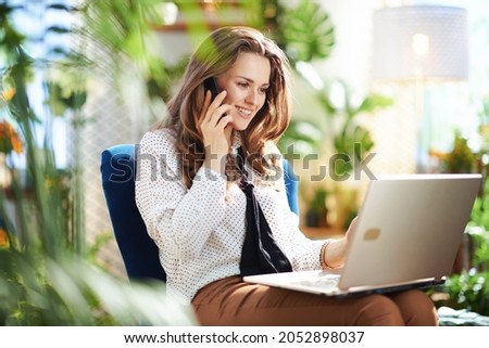 Green Home. happy modern 40 years old woman with long wavy hair in the modern house in sunny day using a smartphone and using laptop while sitting in a blue armchair. Royalty-Free Stock Photo #2052898037