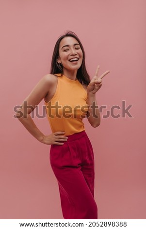 Young asian lady with brown hair smiles at camera and holds victory sign upwards. Pretty brunette model in casual summer outfit posing holding her hip and standing over pink wall.