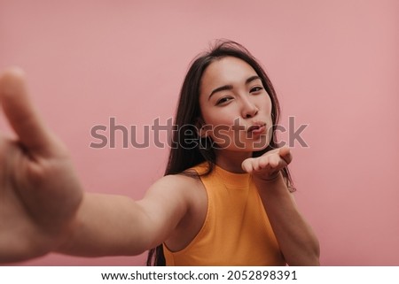 Close-up portrait of beautiful asian girl blowing air kiss for new boyfriend on pink background. Brunette model makes selfie while looking at camera, wearing yellow top. Love and comfort concept