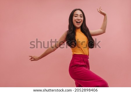 Cheerful asian girl with loose dark hair having fun and posing on pink background. High quality photo of young beauty dancing in studio, in summer colorful clothes. Royalty-Free Stock Photo #2052898406