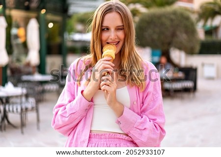 Summer outdoor portrait of pretty blonde woman eating tasty Italian gelato cone ice cream, enjoy her vacation, pink trendy outfit.