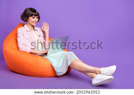 Full length photo of happy pretty young woman sit chair hold laptop wave hello camera isolated on purple color background