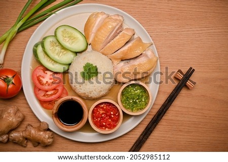 Delicious Hainanese chicken rice is served with three different sauces, as well as cucumber and tomato slides in a plate on wooden table. top view