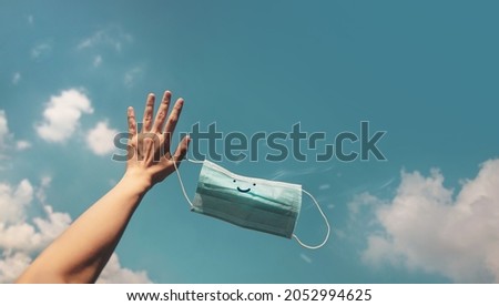 Hope, Positive Mind for Coronavirus Concept. Hand Raised Up a Smiling Medical Mask into the Blue Sky. Gesture means Goodbye. Take-Off Mask and Deep Breathing for Fresh Air Royalty-Free Stock Photo #2052994625