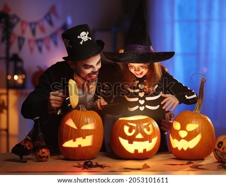 Happy family father and daughter in Halloween costumes looking inside of glowing jack-o-lantern, celebrating all hallows eve together in dark room decorated with carved pumpkins and scary decorations