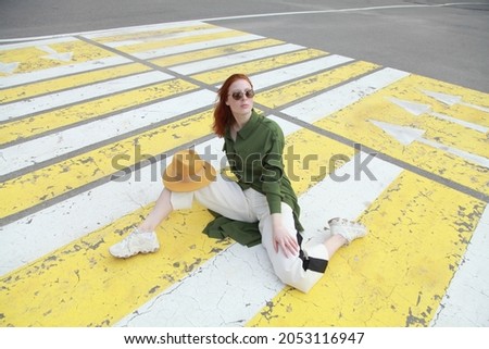 Sporty ginger girl wearing fashion sport chic clothes sitting on street zebra crossing road, outdoor shoot, urban style. Female model in swag clothes posing outside over pedestrian crosswalk.

