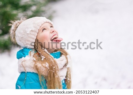 Portrait of a cute blonde with blue eyes in a hat and scarf in winter. A teenager in the forest catches snowflakes with his mouth. Happy child in winter clothes