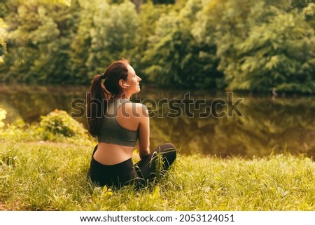 Sporty woman is taking a break for recovery near a lake in lotus pose while listening through wireless earphones.