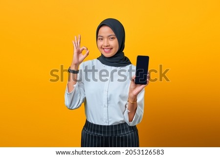 Happy Asian woman showing mobile phone blank screen and hand gesture okay on yellow background
