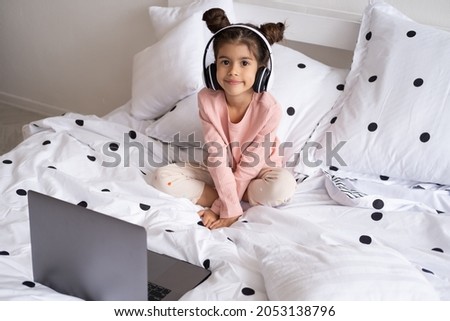 Smiling happy little kid girl using laptop in wireless headphones in bed at home