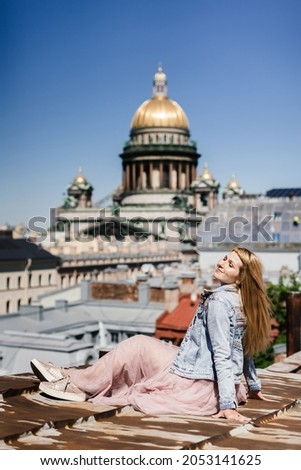 happy young woman sits on the rusty rooftops of the city and enjoys the sunbeams and the view of the tall cathedral. tourist on the saint petersburg rooftops. tourist trend. visiting card of the city.