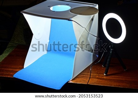 a white plastic lightbox with a blue background and a selfie ring lamp on a tripod stand on a brown table