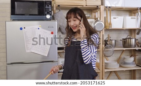 young asian female cooking in kitchen having pleasant phone talk. taiwanese lady wearing apron walking away from stove with wooden spoon in hand. burst out laughing. genuine lifestyle