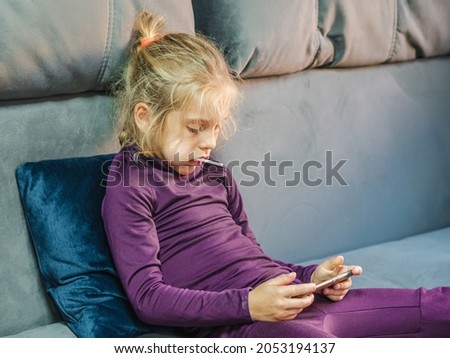 A blond-haired girl with a lollipop in her mouth, watching cartoons on a smartphone on a gray couch. The concept of a digital childhood