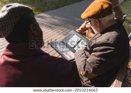 Elderly man holding photo album near blurred interracial friends on bench in park