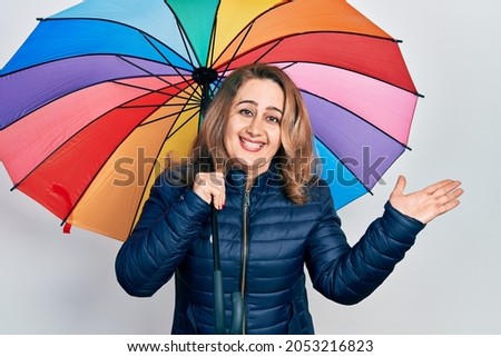 Middle age caucasian woman holding colorful umbrella celebrating achievement with happy smile and winner expression with raised hand 