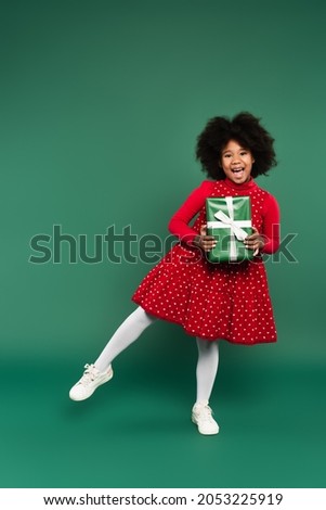 Excited african american kid in red dress holding present on green background