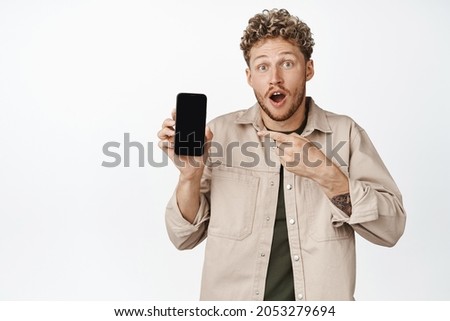 Excited blond curly guy pointing finger at mobile phone screen, looking surprised, showing awesome news online, standing over white background