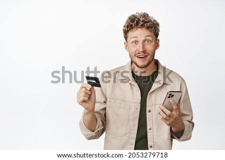 Handsome blond curly guy holding smartphone and credit card, looking excited about store sale, paying online, order in mobile phone, white background