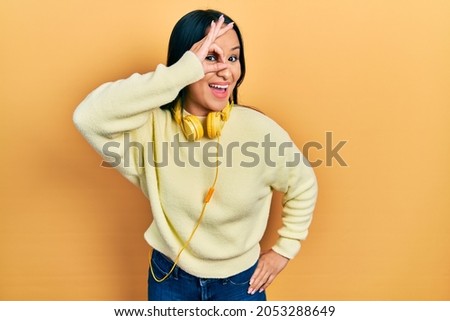Beautiful hispanic woman with nose piercing wearing headphones on neck smiling happy doing ok sign with hand on eye looking through fingers 