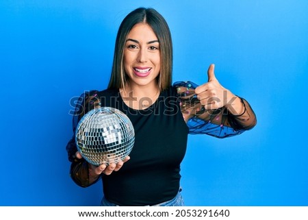 Young hispanic woman holding shiny disco ball smiling happy and positive, thumb up doing excellent and approval sign 