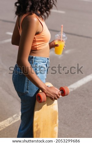 cropped view of curly african american woman holding plastic cup with orange juice and longboard outside