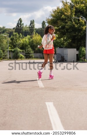 full length of smiling african american woman skating on roller skates while holding plastic cup and smartphone