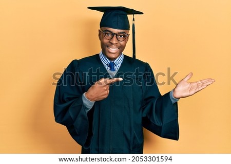 Young african american man wearing graduation cap and ceremony robe amazed and smiling to the camera while presenting with hand and pointing with finger. 