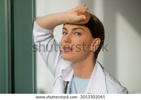A dark-haired doctor standing near the window and looking thoughtful